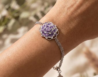 Puede incluir: Una pulsera plateada con un diseño floral morado detallado. La flor es el punto focal, con un efecto de pétalos en capas. La pulsera tiene una cadena delicada y un pequeño dije redondo con texto.