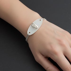 May include: Silver bracelet featuring an oval white charm with a detailed silver car design. The bracelet has a delicate chain and is displayed on a wrist against a dark background. A stylish accessory.