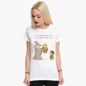 May include: White t-shirt featuring a cartoon illustration of characters from The Lord of the Rings. The image shows Gandalf offering something to Frodo and Samwise with the text "IT'S DANGEROUS TO GO ALONE. TAKE THIS."