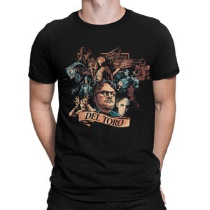 May include: Black t-shirt featuring a graphic design with multiple figures and the text "DEL TORO" on a banner. The design includes images of various characters and faces, with a dark color palette.
