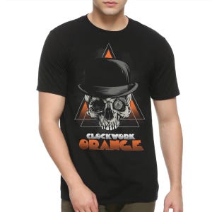 Può includere: T-shirt nera con un teschio con bombetta, su un triangolo arancione, e le parole "CLOCKWORK ORANGE" in arancione e bianco. Il disegno è centrato sul petto.