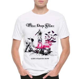 May include: White t-shirt with a graphic design. The design features the text "Three Days Grace" in a gothic font, figures with axes, a burst of pink and black, and the words "LIFE STARTS NOW".