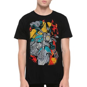 May include: A black short-sleeved t-shirt featuring a graphic print of a yellow and blue costumed character with metal claws riding a teal motorcycle. The design includes stylised red and orange flames in the background behind the character.