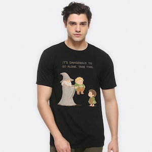 Op de afbeelding: Zwart T-shirt met een cartoon illustratie van Gandalf die een cadeau aan Frodo en Sam Gamgee geeft. De tekst "IT'S DANGEROUS TO GO ALONE. TAKE THIS." staat boven de karakters. Het shirt is gemaakt van zacht materiaal.