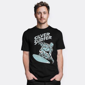 May include: Black t-shirt with a comic book style illustration of the Silver Surfer surfing on a light blue surfboard. The words "Silver Surfer" are printed above the illustration in white.