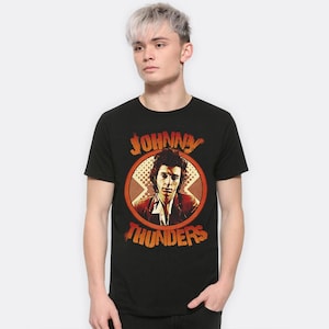 Puede incluir: Camiseta negra con un retrato de Johnny Thunders de estilo vintage en un diseño circular. El diseño incluye el nombre "JOHNNY THUNDERS" en letras naranjas. El retrato tiene un esquema de color rojo y naranja.