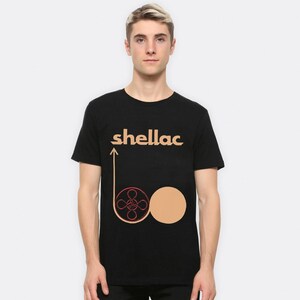 May include: Black t-shirt with the word "shellac" in tan letters above a graphic of a reel and a circle in tan and red. The shirt is a crew neck and short-sleeved.
