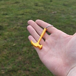 May include: A small, bright yellow plastic hammer keychain featuring a claw hammer head and a straight handle. The tool is attached to a circular silver-toned metal split ring, providing a functional way to secure keys or attach to bags and accessories.