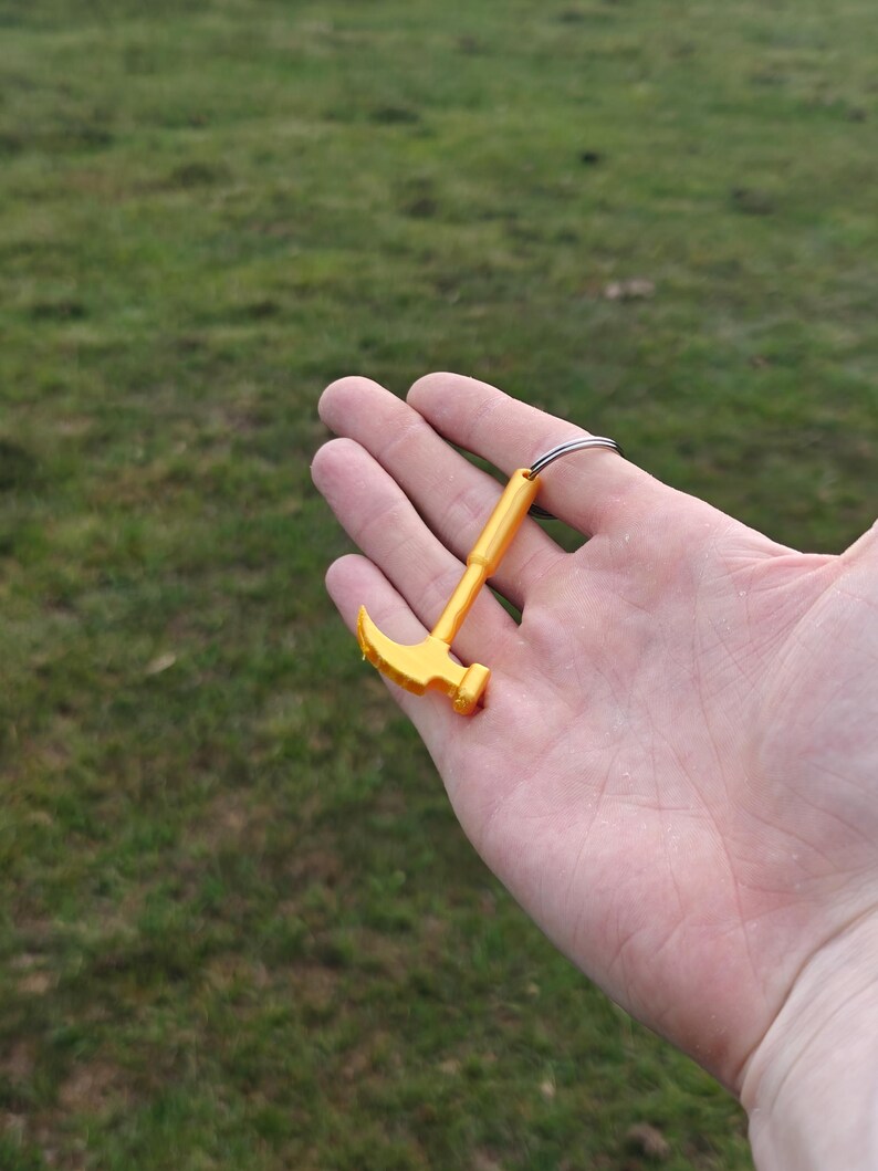 May include: A small, bright yellow plastic hammer keychain featuring a claw hammer head and a straight handle. The tool is attached to a circular silver-toned metal split ring, providing a functional way to secure keys or attach to bags and accessories.