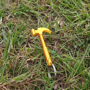 May include: A bright yellow metal hammer keychain featuring a classic claw hammer head design attached to a circular silver metal split ring. The tool is constructed with a smooth, solid handle and a curved claw, providing a functional miniature representation of a standard construction hammer.