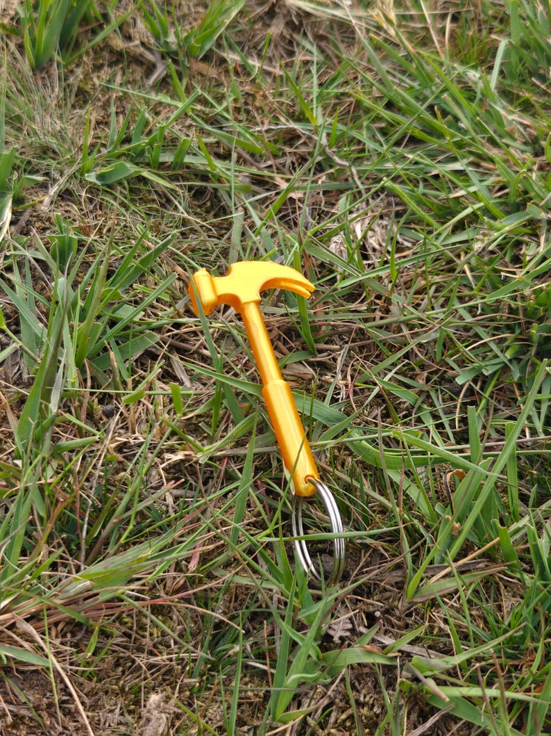 May include: A bright yellow metal hammer keychain featuring a classic claw hammer head design attached to a circular silver metal split ring. The tool is constructed with a smooth, solid handle and a curved claw, providing a functional miniature representation of a standard construction hammer.