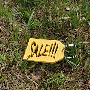 May include: A yellow, tag-shaped keychain with the word SALE!!! cut out from the center. The tag has a textured surface and is attached to a circular silver metal split ring. The design is flat and features bold, stylized lettering.