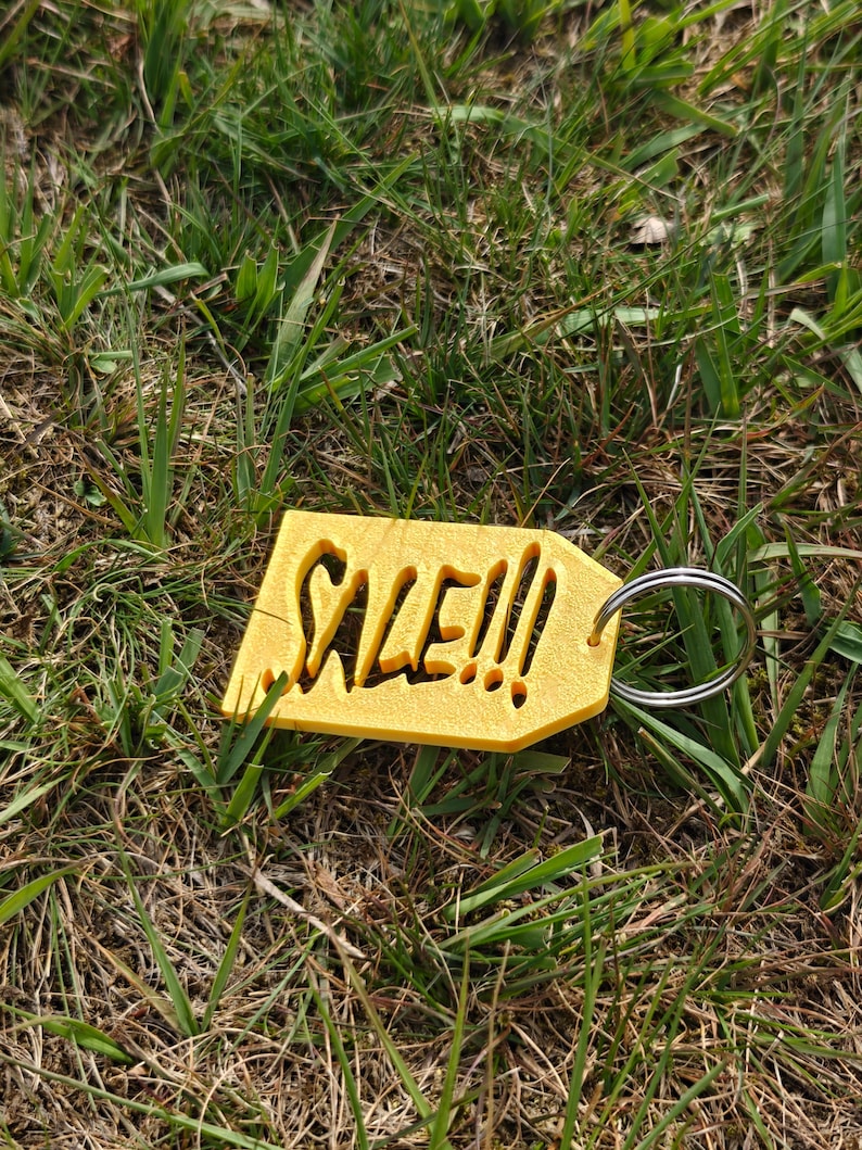 May include: A yellow, tag-shaped keychain with the word SALE!!! cut out from the center. The tag has a textured surface and is attached to a circular silver metal split ring. The design is flat and features bold, stylized lettering.