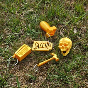 May include: A collection of four gold-colored keychains including a bomb, a skull, a hammer, and a rectangular block with the text 250 CAL. 50 CARTRIDGES UNITED BALL M1909 LOT LCL-15279, alongside a gold-colored tag with the text SALE!!!.