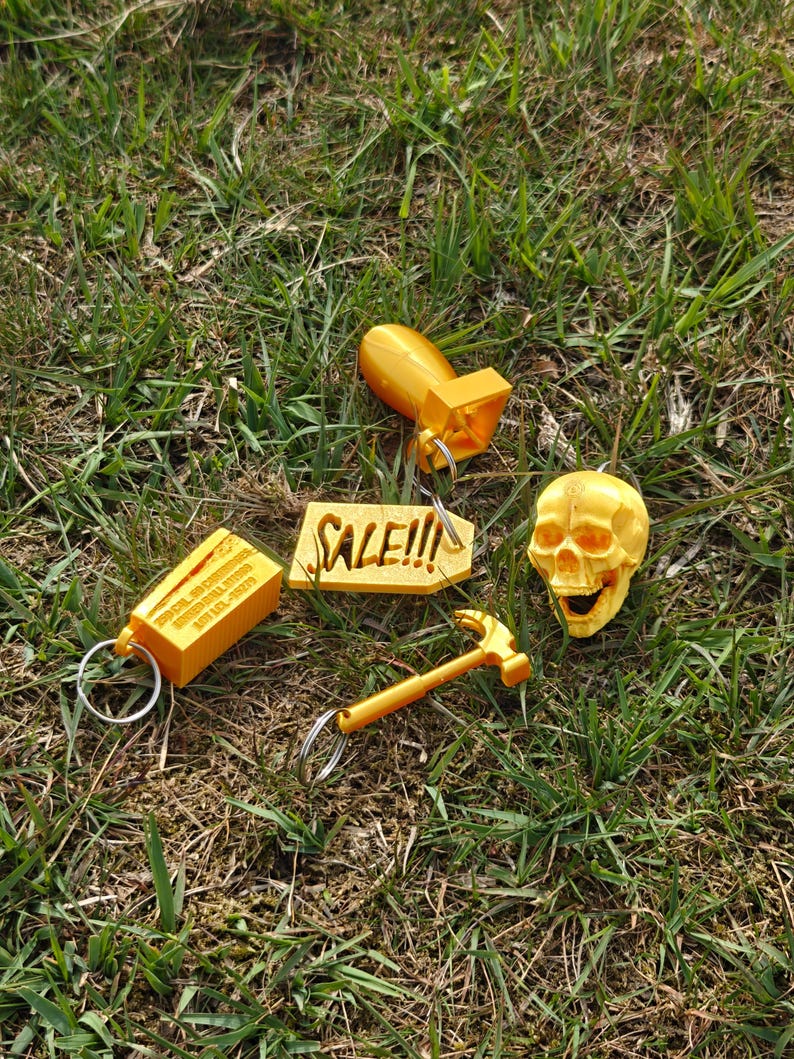 May include: A collection of four gold-colored keychains including a bomb, a skull, a hammer, and a rectangular block with the text 250 CAL. 50 CARTRIDGES UNITED BALL M1909 LOT LCL-15279, alongside a gold-colored tag with the text SALE!!!.