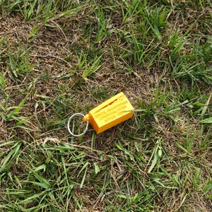 May include: A bright yellow rectangular keychain with a silver metal ring. The keychain features embossed text that reads 250 CAL .50 CARTRIDGES LINKED BALL M1989 LOT LCL-15279, along with a small graphic of a cartridge.