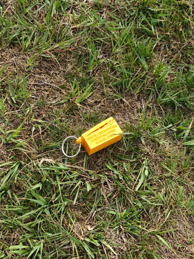 May include: A bright yellow rectangular keychain with a silver metal ring. The keychain features embossed text that reads 250 CAL .50 CARTRIDGES LINKED BALL M1989 LOT LCL-15279, along with a small graphic of a cartridge.