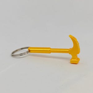 May include: A bright yellow keychain shaped like a miniature claw hammer. The tool-themed charm features a smooth, solid handle and a curved claw head, attached to a standard circular silver-colored metal split ring for keys.
