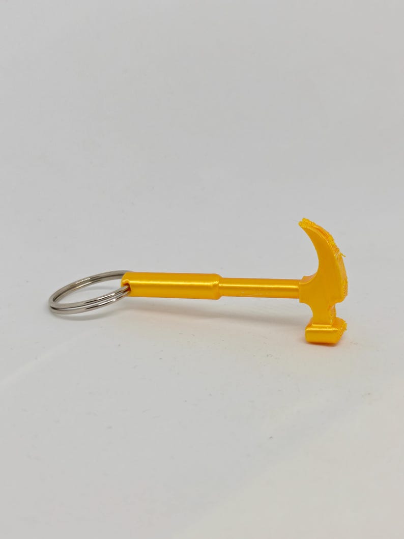 May include: A bright yellow keychain shaped like a miniature claw hammer. The tool-themed charm features a smooth, solid handle and a curved claw head, attached to a standard circular silver-colored metal split ring for keys.
