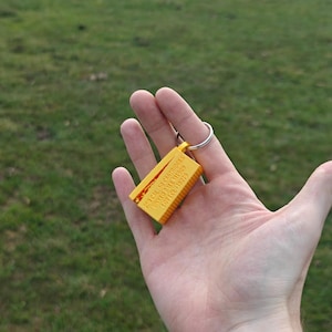 May include: A yellow rectangular keychain featuring a ribbed texture and embossed text that reads 250 CAL 50 CARTRIDGES LINKED BALL M1909 LOT LCE-15279. The object is attached to a circular silver metal split ring for keys.