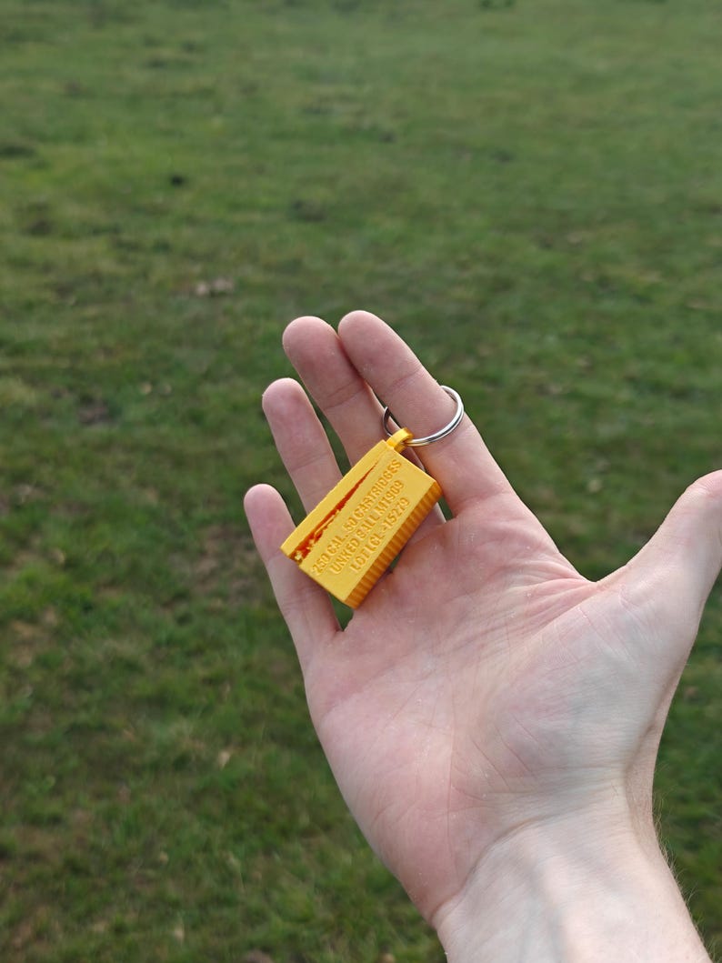 May include: A yellow rectangular keychain featuring a ribbed texture and embossed text that reads 250 CAL 50 CARTRIDGES LINKED BALL M1909 LOT LCE-15279. The object is attached to a circular silver metal split ring for keys.