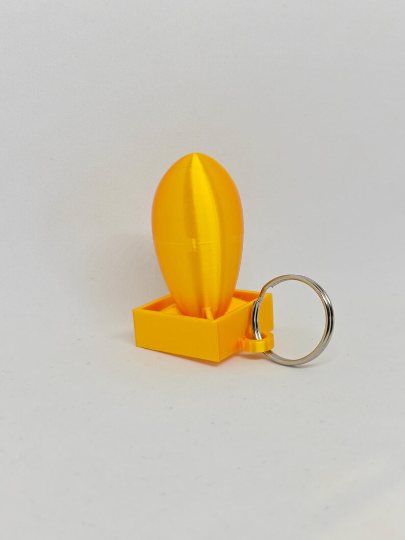 May include: A bright yellow 3D printed keychain featuring a smooth, vertically oriented oval shape attached to a square base. A silver metal split ring is connected to the side of the square base, allowing for attachment to keys or other items.