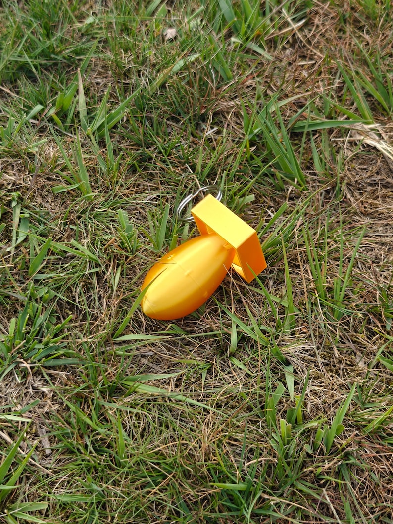 May include: A bright yellow, bomb-shaped keychain featuring a smooth, rounded body and a square-shaped fin assembly at the rear. The object is constructed from a solid, molded material and includes a standard silver-colored metal split ring for attachment.
