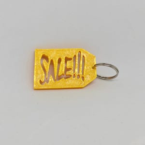 May include: A yellow, rectangular tag-shaped keychain with a textured, glittery finish. The word SALE!!! is cut out from the center of the tag in a bold, stylized font. A silver-colored metal split ring is attached to the right side of the tag.