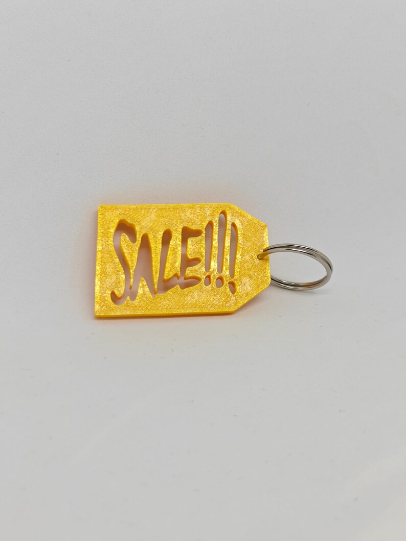 May include: A yellow, rectangular tag-shaped keychain with a textured, glittery finish. The word SALE!!! is cut out from the center of the tag in a bold, stylized font. A silver-colored metal split ring is attached to the right side of the tag.