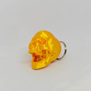May include: A bright yellow, three-dimensional skull keychain featuring a realistic anatomical design with an open jaw. The skull has a smooth, glossy finish and is attached to a standard circular silver-colored metal split ring for keys.