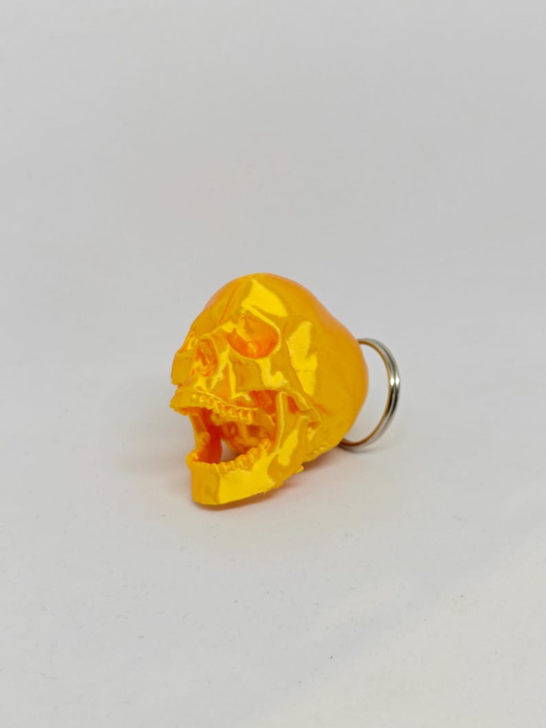 May include: A bright yellow, three-dimensional skull keychain featuring a realistic anatomical design with an open jaw. The skull has a smooth, glossy finish and is attached to a standard circular silver-colored metal split ring for keys.