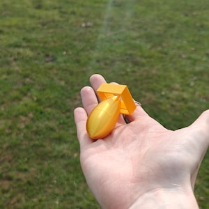 May include: A small, bright yellow, three-dimensional printed object shaped like a stylized bomb with a smooth, rounded body and a square-shaped fin assembly at the rear, featuring a metallic sheen and a compact, handheld size.