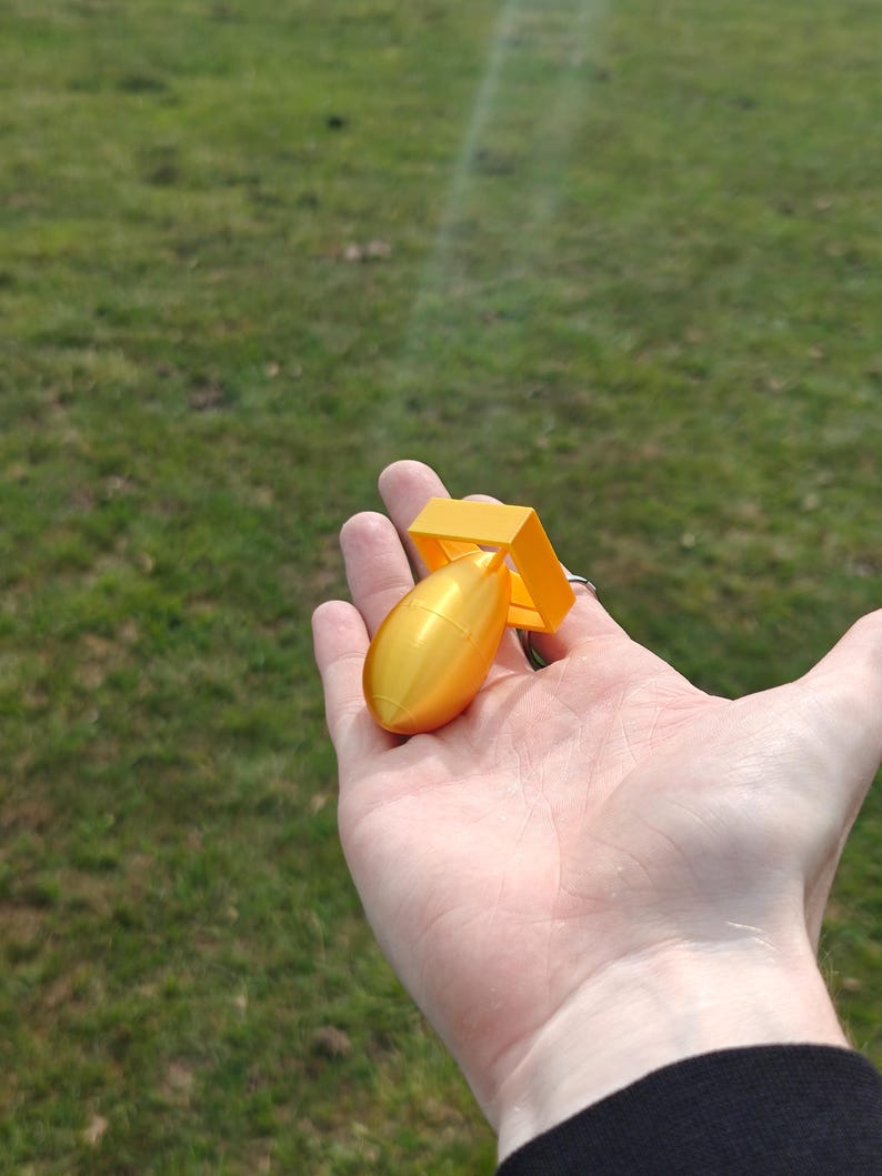 May include: A small, bright yellow, three-dimensional printed object shaped like a stylized bomb with a smooth, rounded body and a square-shaped fin assembly at the rear, featuring a metallic sheen and a compact, handheld size.
