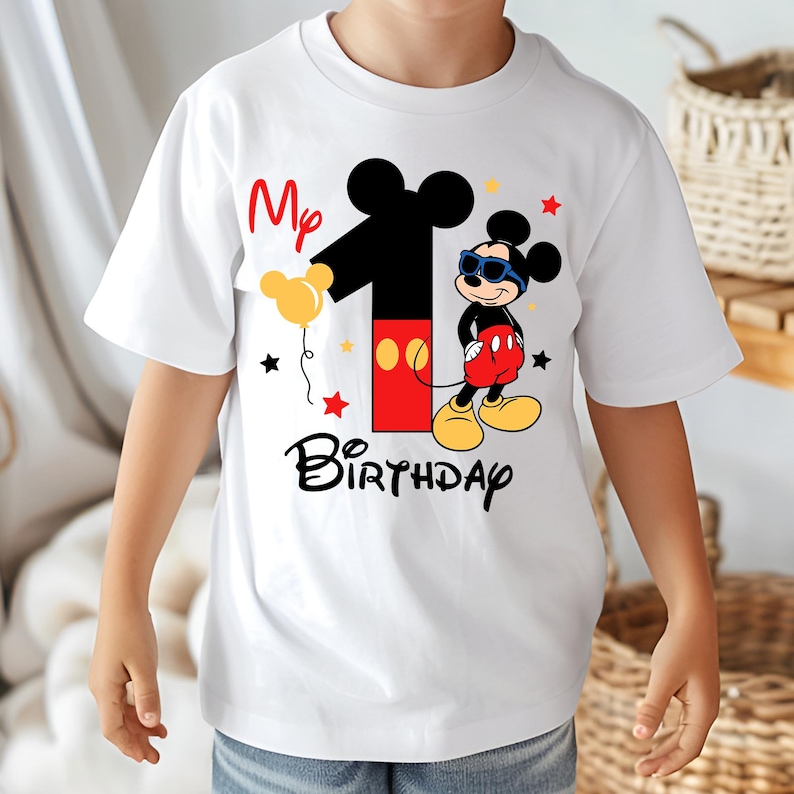 May include: White t-shirt featuring a Mickey Mouse design for a first birthday. The design includes a large number 1, Mickey Mouse wearing sunglasses, the words "Birthday", and the text "My". The shirt has short sleeves.