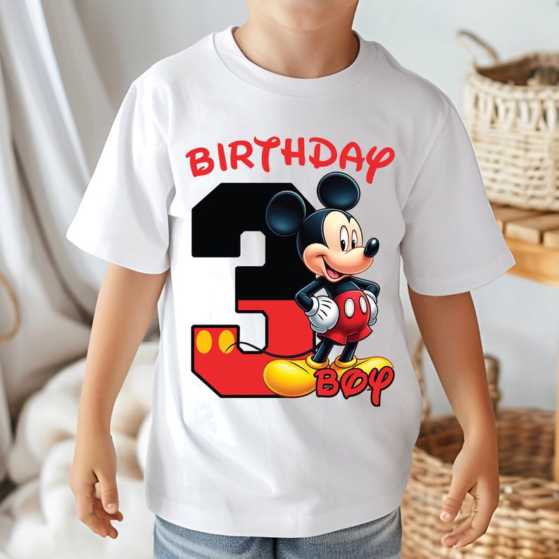 May include: White t-shirt featuring a colorful graphic of Mickey Mouse and the number 3. The text "BIRTHDAY BOY" is printed in red. The shirt is designed for a child's third birthday.