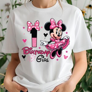 May include: White t-shirt featuring a graphic of Minnie Mouse in a pink polka-dot dress and bow. The design includes the number "1" and the text "Birthday Girl" in pink. Pink stars and mouse silhouettes complete the design.