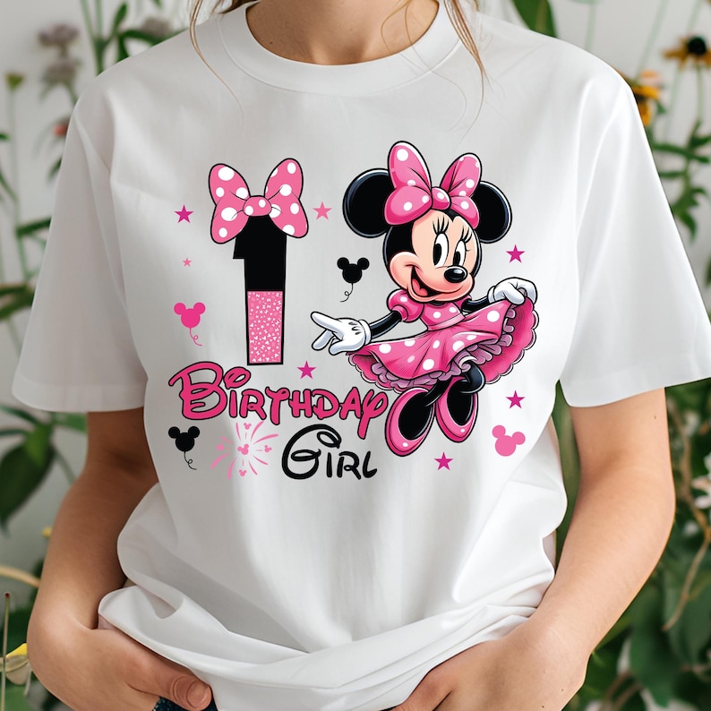 May include: White t-shirt featuring a graphic of Minnie Mouse in a pink polka-dot dress and bow. The design includes the number "1" and the text "Birthday Girl" in pink. Pink stars and mouse silhouettes complete the design.