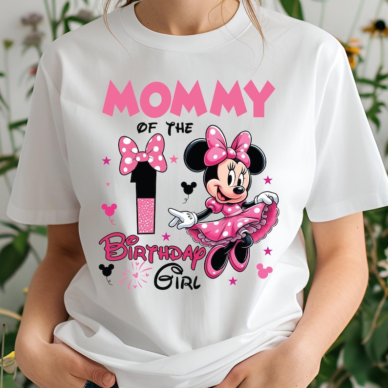 May include: White t-shirt featuring a Minnie Mouse design. The shirt has the text "MOMMY OF THE 1 BIRTHDAY GIRL" in pink. Minnie Mouse is wearing a pink dress and bow. The design includes stars and Mickey Mouse silhouettes.