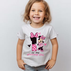 K&ouml;nnte beinhalten: Wei&szlig;es T-Shirt mit einem Minnie-Maus-Design f&uuml;r den ersten Geburtstag. Das Design umfasst die Zahl 1 mit rosa Punkten, eine Schleife und den Text "OF THE BIRTHDAY GIRL".