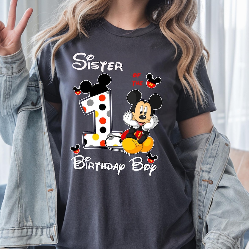 Puede incluir: Camiseta gris oscuro con Mickey Mouse y el texto "Sister of the Birthday Boy". El n&uacute;mero "1" est&aacute; decorado con lunares rojos, amarillos y blancos. Mickey Mouse lleva una chaqueta roja y zapatos amarillos.