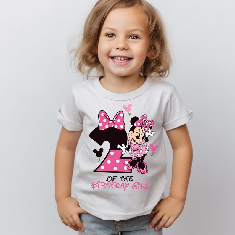 K&ouml;nnte beinhalten: Wei&szlig;es T-Shirt mit einem Minnie Mouse-Design, der Zahl 2, Punkten und dem Text "OF THE BIRTHDAY GIRL". Das Design beinhaltet eine gro&szlig;e Schleife und Minnie Mouse in einem rosa gepunkteten Kleid. Das T-Shirt ist f&uuml;r ein Kind.