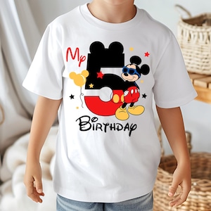 May include: White t-shirt featuring a graphic of Mickey Mouse with sunglasses, the number 5, and the words "My Birthday." The design includes red, black, yellow, and blue elements, with star accents. The shirt is for a child.