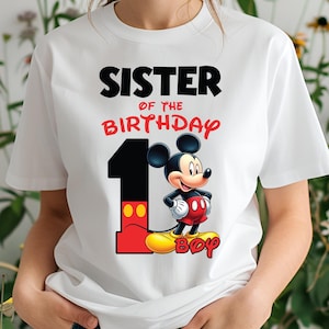 Op de afbeelding: Wit T-shirt met de tekst "SISTER OF THE BIRTHDAY" in zwart en rood. Een groot nummer "1" staat naast een cartoonafbeelding van Mickey Mouse. Het woord "BOY" staat onder Mickey Mouse.