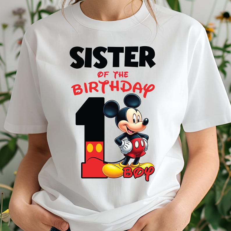 Op de afbeelding: Wit T-shirt met de tekst "SISTER OF THE BIRTHDAY" in zwart en rood. Een groot nummer "1" staat naast een cartoonafbeelding van Mickey Mouse. Het woord "BOY" staat onder Mickey Mouse.