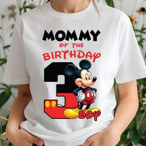 May include: White t-shirt featuring the text "MOMMY OF THE BIRTHDAY BOY" in black and red. A large number 3 is in black and red, with a cartoon Mickey Mouse character. The shirt is a crew neck style.