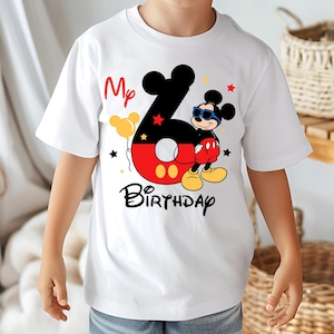 May include: White t-shirt featuring a colorful Mickey Mouse design for a sixth birthday. The design includes the number 6, Mickey Mouse wearing sunglasses, the words "My Birthday", and star and balloon graphics. A fun and festive shirt.
