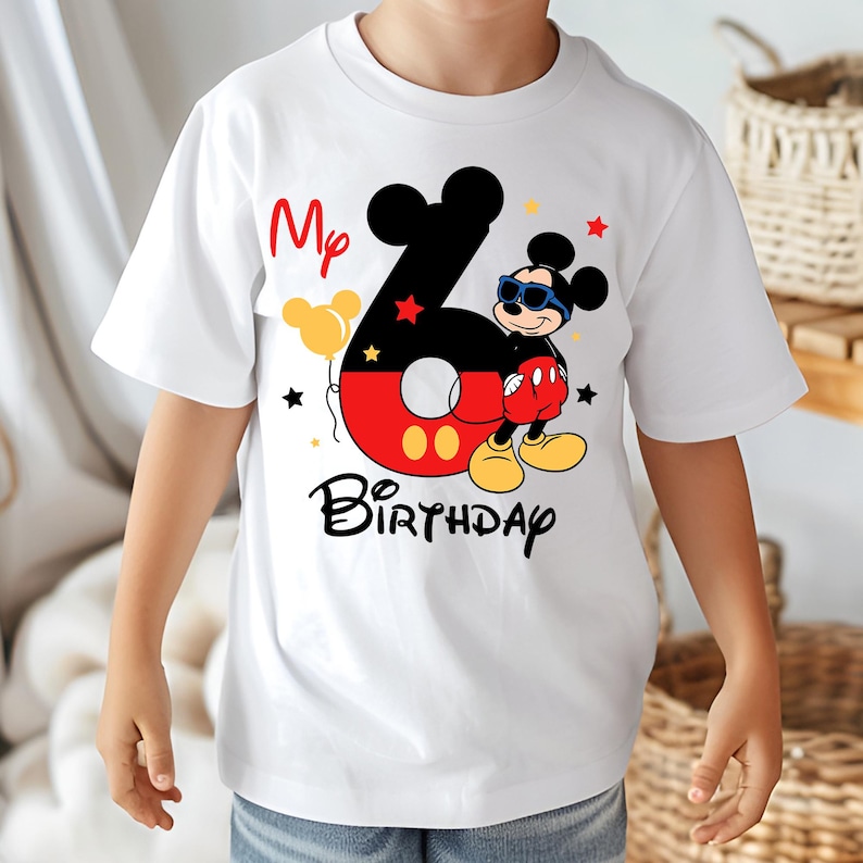 May include: White t-shirt featuring a colorful Mickey Mouse design for a sixth birthday. The design includes the number 6, Mickey Mouse wearing sunglasses, the words "My Birthday", and star and balloon graphics. A fun and festive shirt.