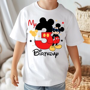 May include: White t-shirt featuring a Mickey Mouse design for a third birthday. The design includes a large red number 3, the words "My Birthday", and a cartoon Mickey Mouse wearing sunglasses. Yellow and black accents complete the design.