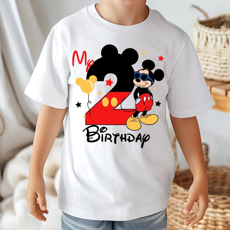 May include: White t-shirt featuring a colorful Mickey Mouse design for a second birthday. The design includes a large number 2, Mickey Mouse in sunglasses, the words "My Birthday", and star and balloon accents. A fun and festive shirt.