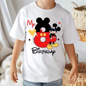 May include: White t-shirt featuring a colorful Mickey Mouse design. The design includes the text "My 8th Birthday" with Mickey Mouse wearing sunglasses. The number 8 is red with yellow balloons and black stars.